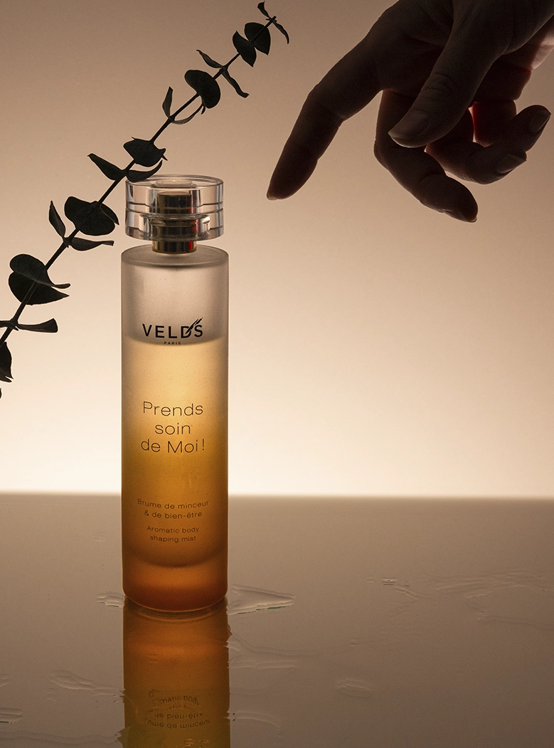 Parfum Velds Prends Soin de Moi, flacon doré, main féminine, eucalyptus séché, lumière dorée chaude