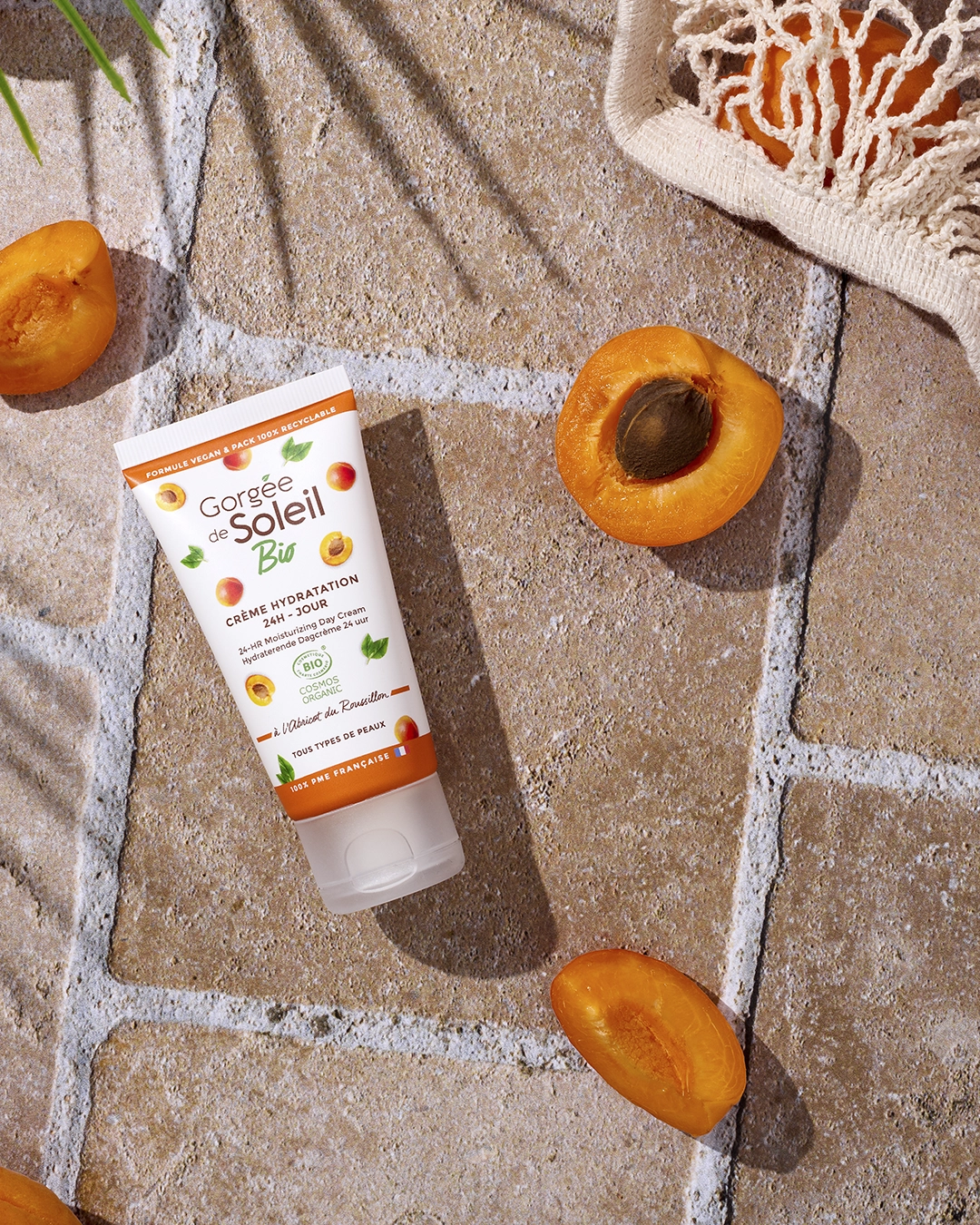 Crème hydratante bio Gorgée de Soleil entourée d’abricots frais sur dalle beige, ambiance estivale