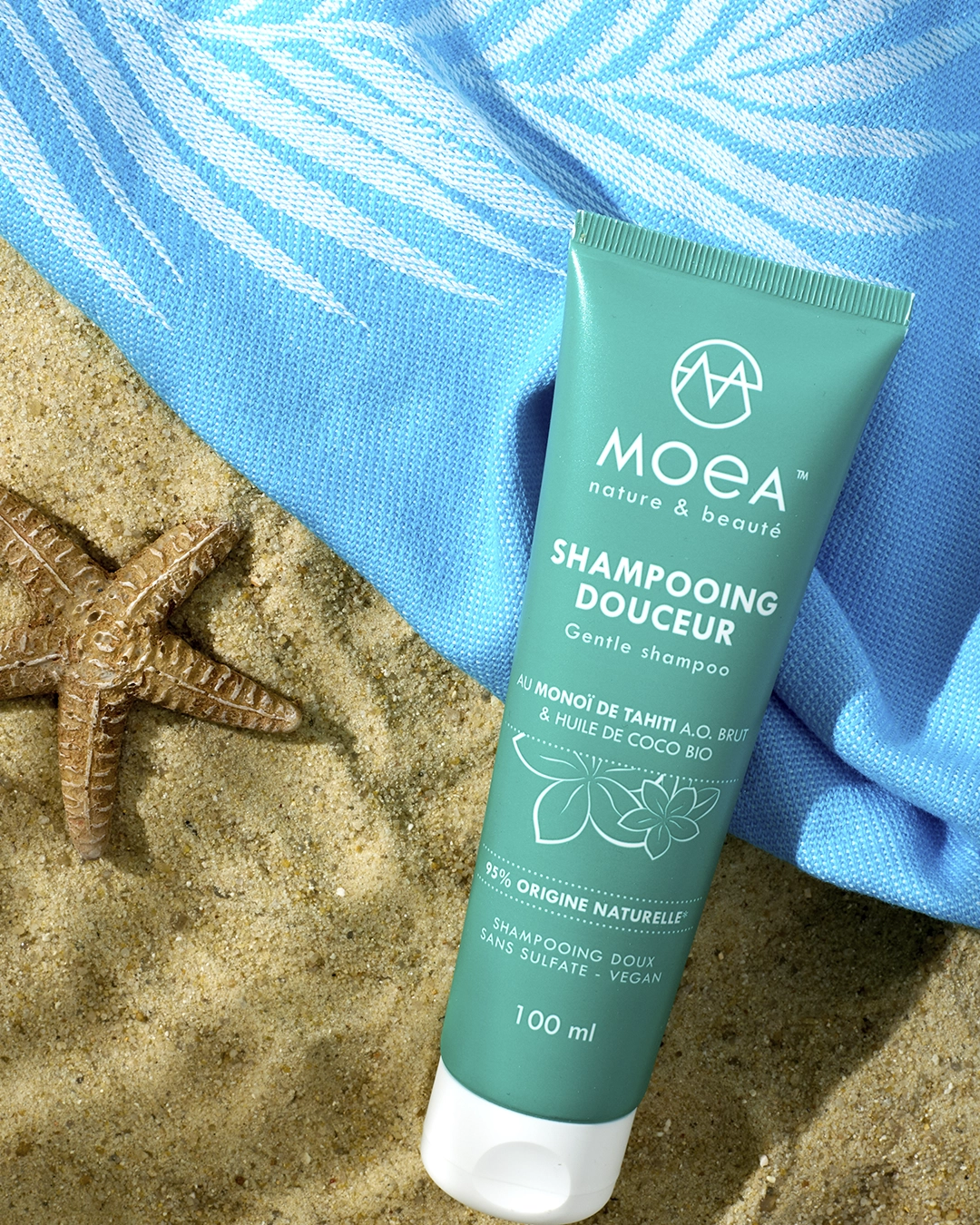 Shampooing douceur Moea posé sur sable, étoile de mer, serviette bleu, ambiance beauté naturelle été
