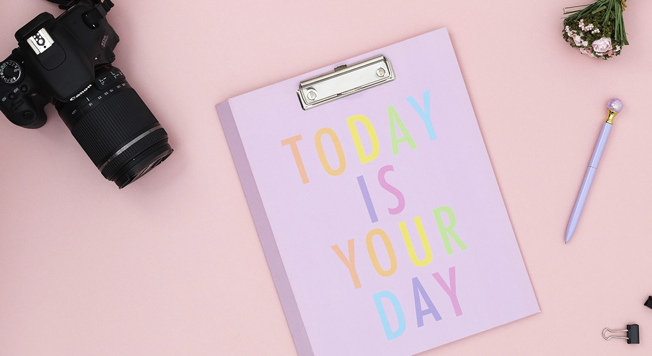 Appareil photo Canon, carnet “Today is your day” et accessoires sur fond rose pastel.