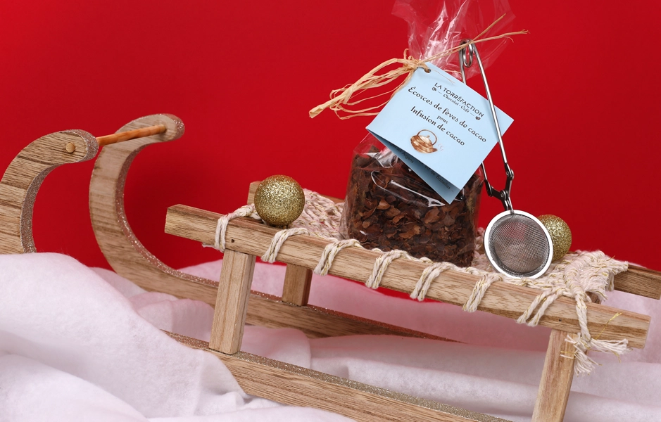 Produit infusion cacao, sachet transparent, traîneau bois, déco boules or, ambiance fête hiver.