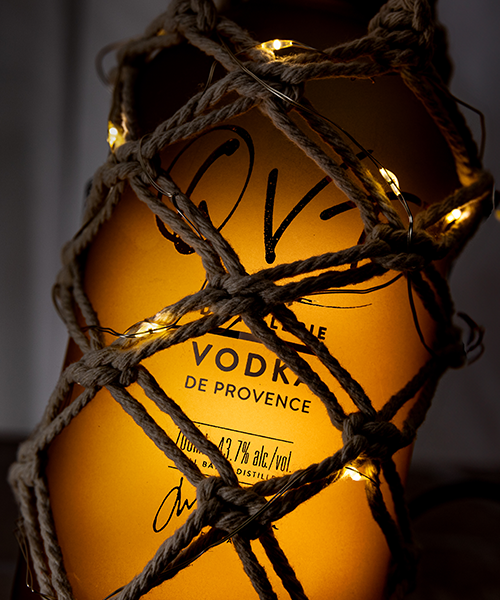 Photo bouteille Vodka QVT Provence éclairée, décor macramé et guirlande lumineuse, ambiance premium