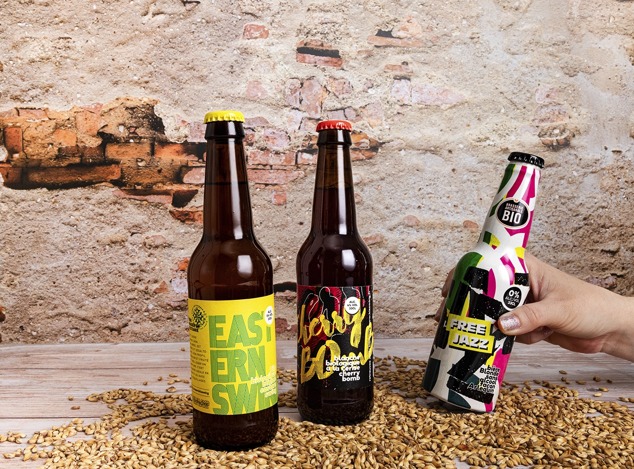 Trois bouteilles de bière artisanale sur grains de malt, main tenant une bouteille colorée.