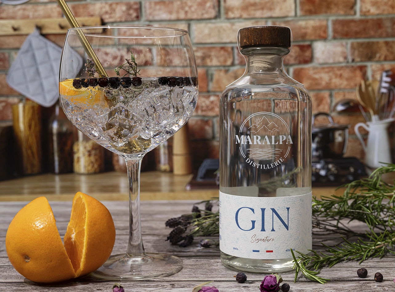 Cocktail gin tonic avec bouteille Maralpa, orange fraîche et aromates sur table en bois.