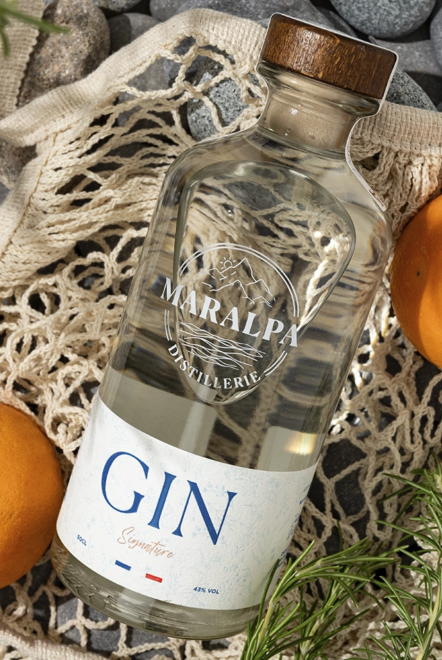 Bouteille gin Maralpa sur galets avec sac filet, orange et romarin frais.