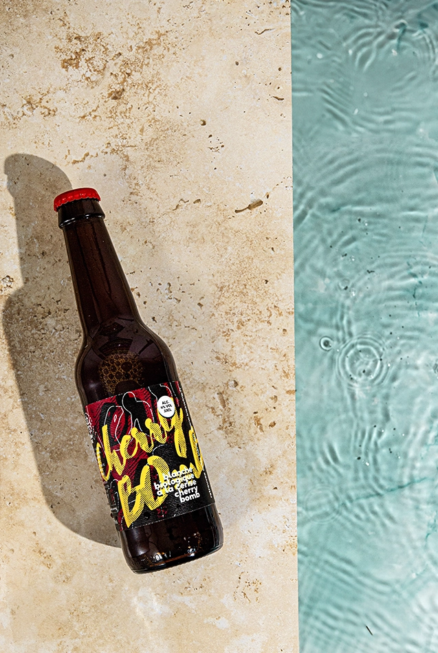 Bouteille de bière Cherry Bomb posée sur pierre claire au bord d’une eau turquoise.