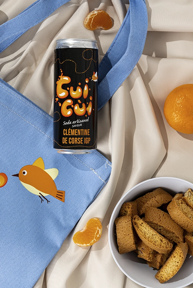 Soda Cui Cui clémentine de Corse avec quartiers d'orange et biscuits sur tissu bleu.