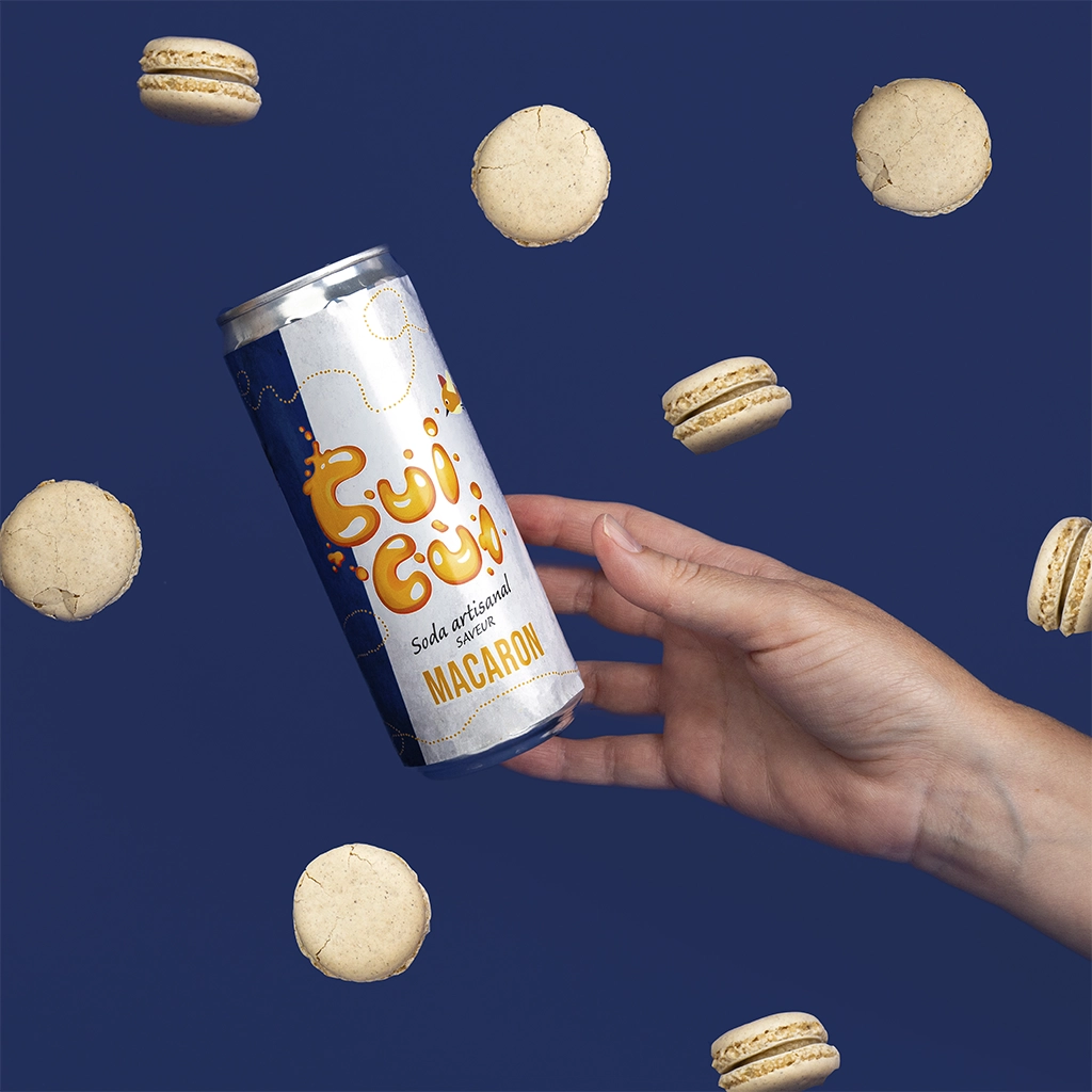 Main tenant canette soda Cui Cui macaron avec macarons vanille flottants sur fond bleu.