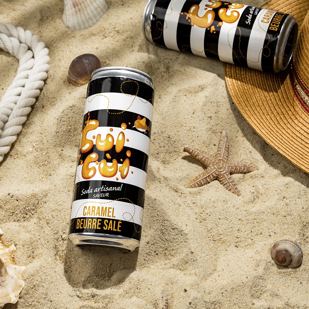 Soda Cui Cui caramel beurre salé sur sable avec coquillages et chapeau de paille.